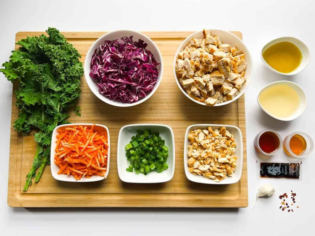 kale, red cabbage, chopped chicken, carrots, green onions, cashews on wooden cutting board and oil, vinegar, honey, soy sauce, garlic, and red pepper flakes next to board