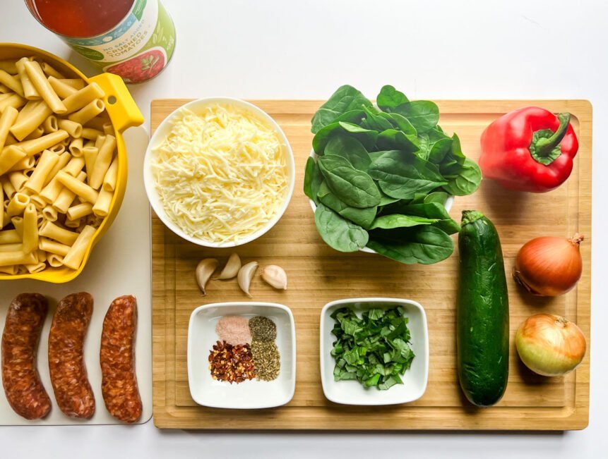 tomato sauce, pasta, italian sausage, cheese, garlic, spices, spinach, basil, zucchini. onions, red bell pepper on a cutting board