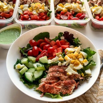 Green goddess salad with mixed greens, tomatoes, carrots, hard-boiled egg, chicken, and cucmbers in bowl with containers in background.