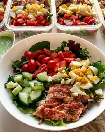 Green goddess salad with mixed greens, tomatoes, carrots, hard-boiled egg, chicken, and cucmbers in bowl with containers in background.