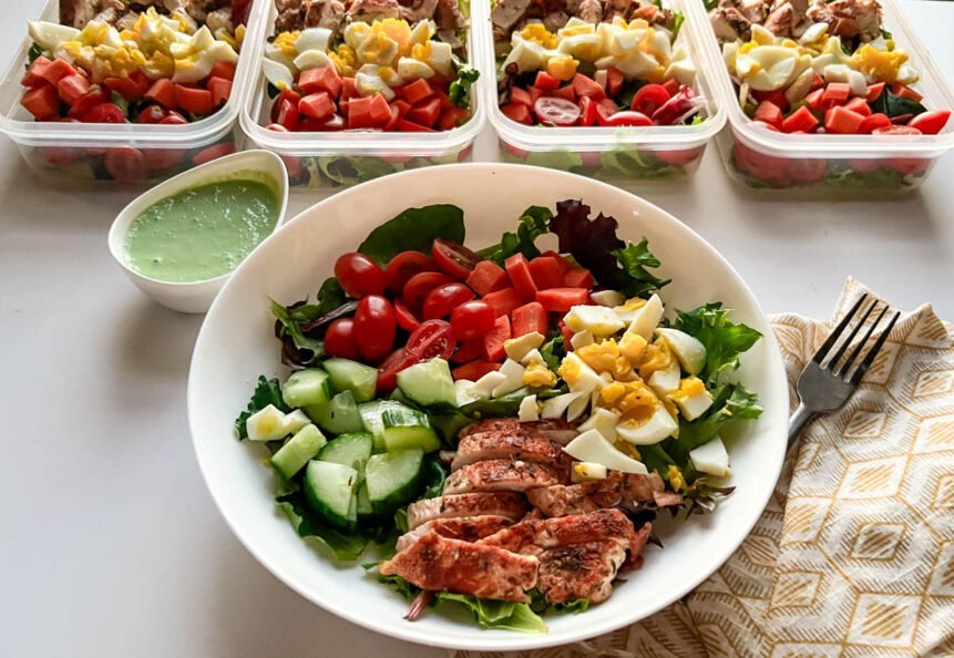 Green goddess salad with mixed greens, tomatoes, carrots, hard-boiled egg, chicken, and cucmbers in bowl with containers in background.