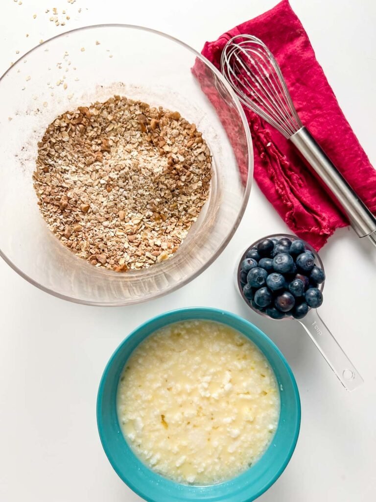 clear bowl with dry ingredients, blue bowl with wet ingredients, blueberries in measuring up, whisk on purple cloth